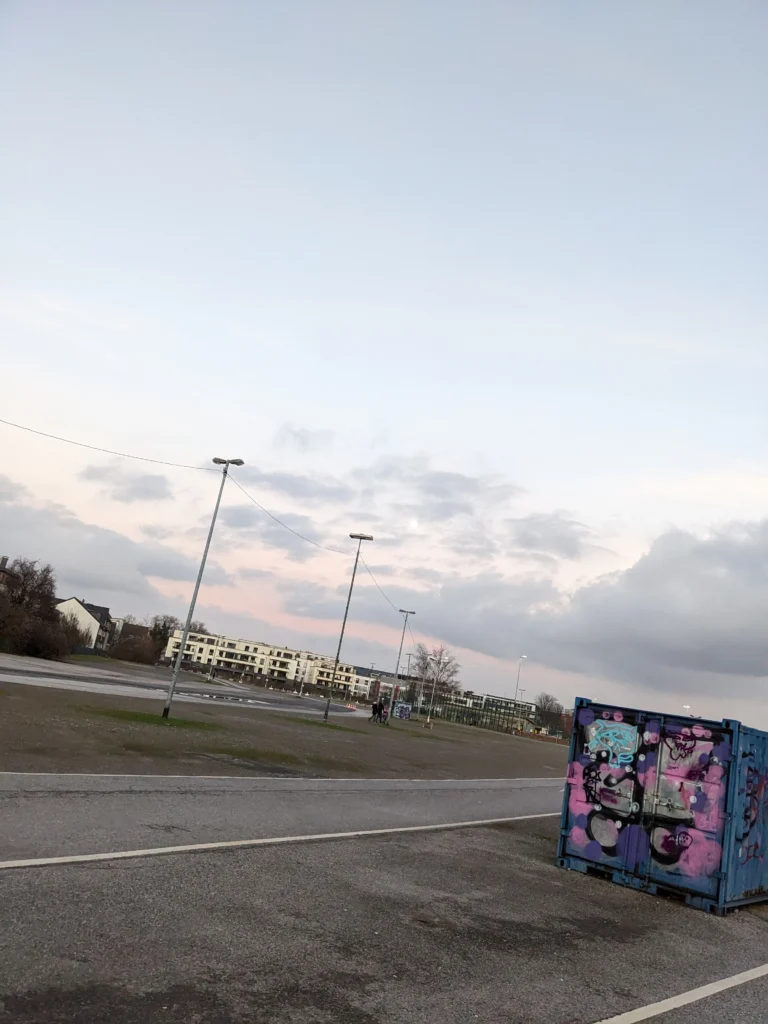 Photo of a parking lot in Essen. It's slightly angled to the left. In the front right is a cargo container with graffiti on it. In the back are houses which form a kind of horizon. The sky is grey to blue with a few clouds and a hint of orange which shows the coming sunset.